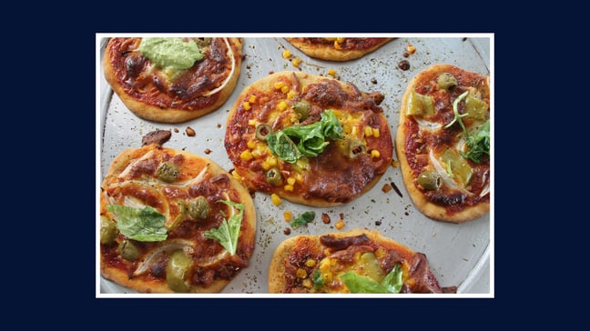 “Tray of homemade mini pizzas topped with tomato sauce, cheese, olives, sweetcorn and herbs.”