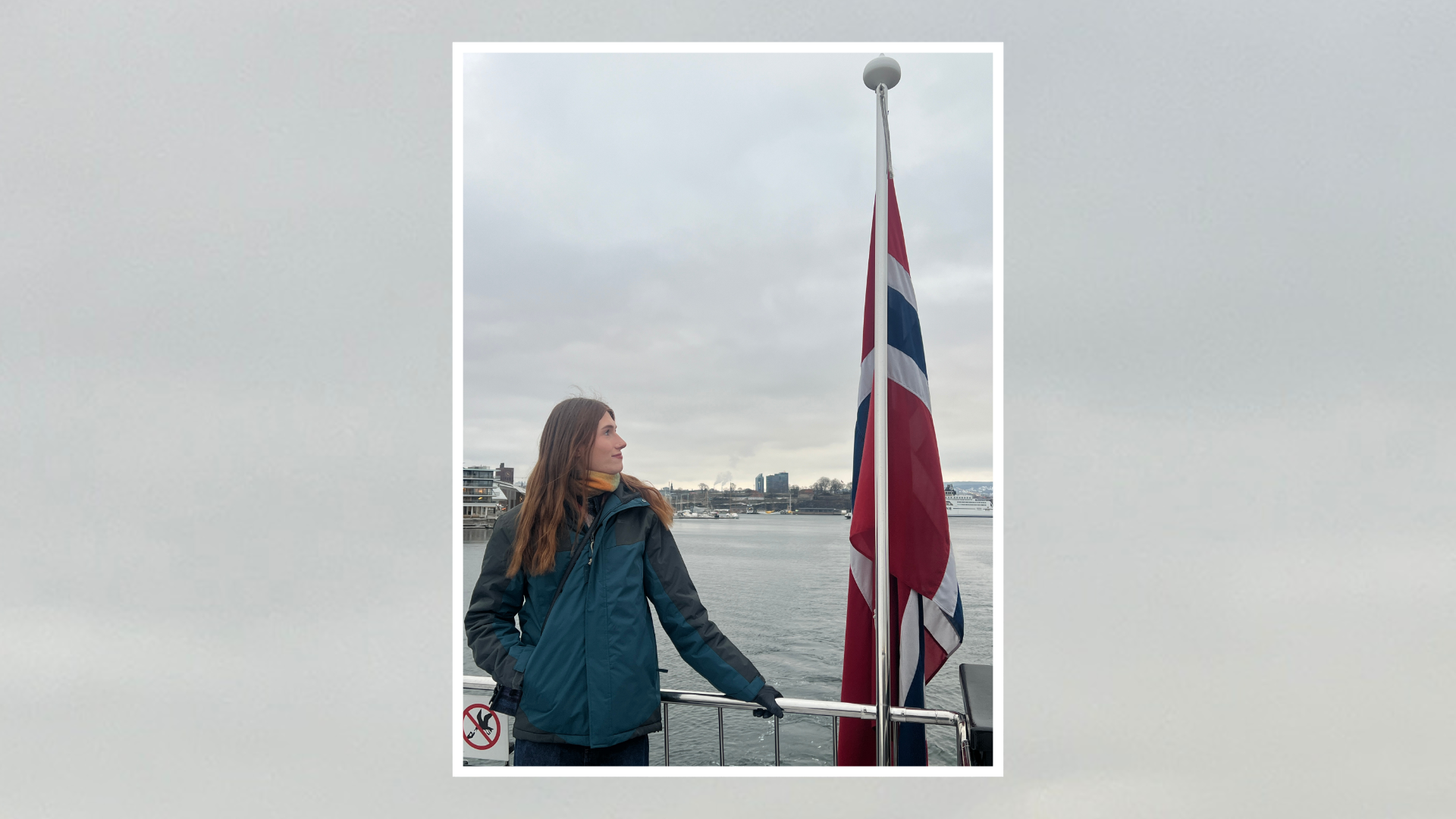Callum stands on the deck of a ferry beside a Norwegian flag, looking across a harbour under a cloudy sky. The photo is framed with a white border on a light grey textured background.