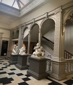 Picture5: the stairwell and entrace hall of the architecture building. A checkquered floor, 3  small white statues and behind them a staircase
