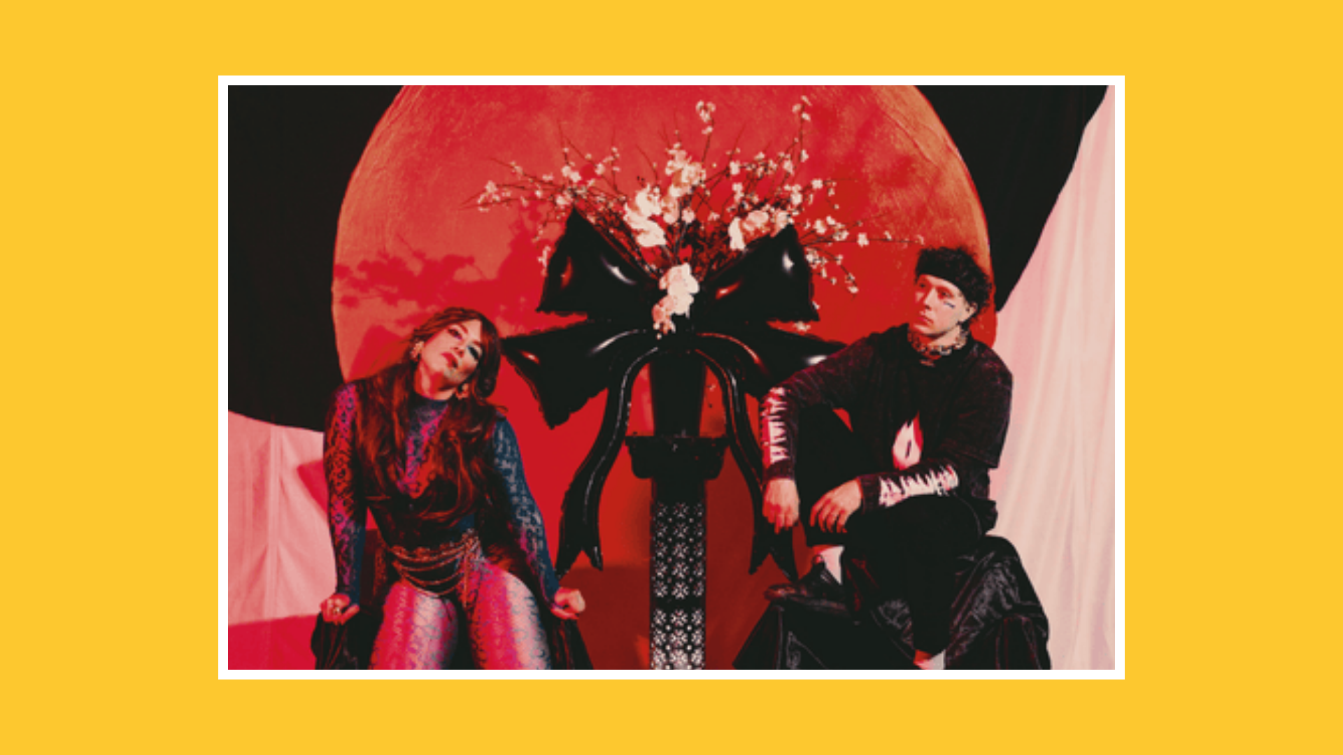 Two performers seated on a stage in front of a large red circular backdrop, styled lighting and theatrical costumes