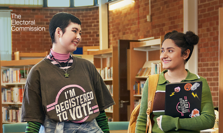 Two students smiling at each other in a library. One wears a T-shirt reading “I’m registered to vote,” while the other holds a laptop with voter registration stickers. The Electoral Commission logo appears in the top left.